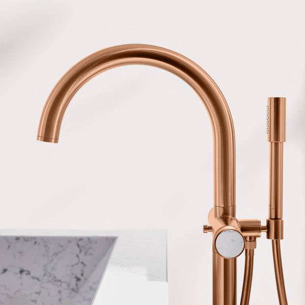 Rose gold freestanding Atrio bathtub faucet with a high-arc spout, side-mounted shower hose, and round control knob, shown beside a marble tub edge on a light background.