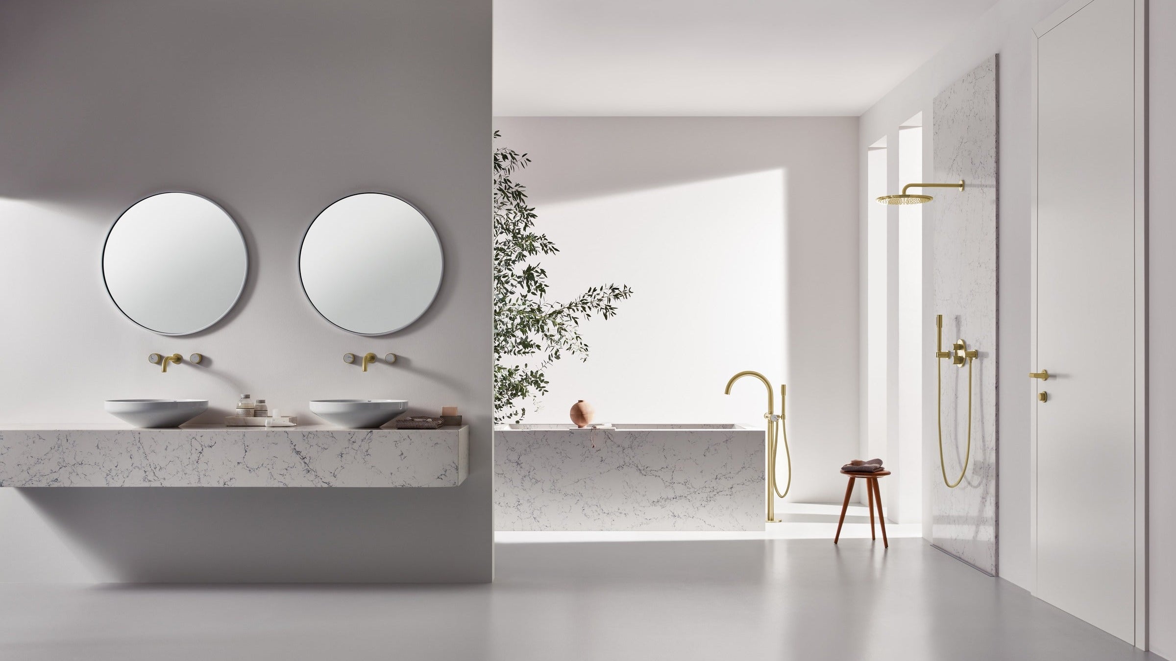 A modern minimalist bathroom interior with a floating double-sink vanity, two round mirrors, and matte brass fixtures. A freestanding bathtub, matching gold shower hardware, and a small wooden stool are visible in the background against white walls.